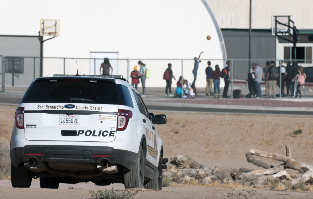 A police vehicle is parked on dirt with children playing basketball in the background.